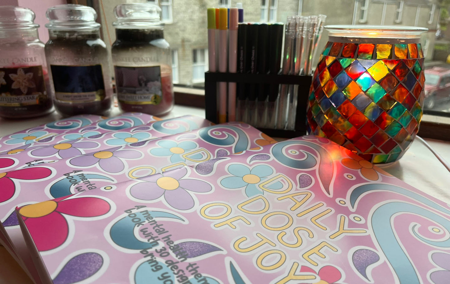 five copies of the daily dose of joy colouring book next to candles and pens